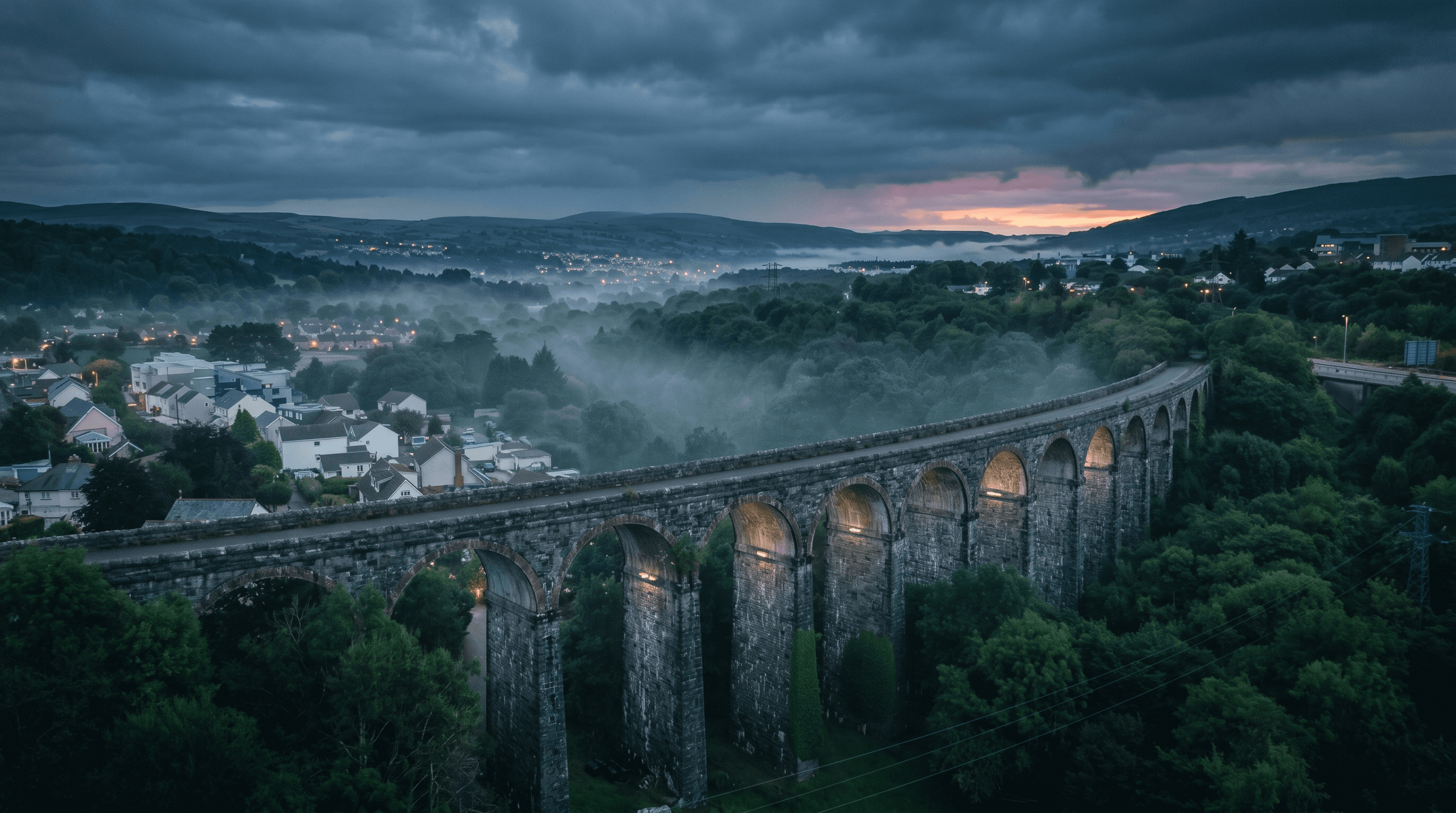 A stone railway viaduct sweeping across a misty Welsh valley town at dusk, dramatic clouds and a pink sunset on the horizon