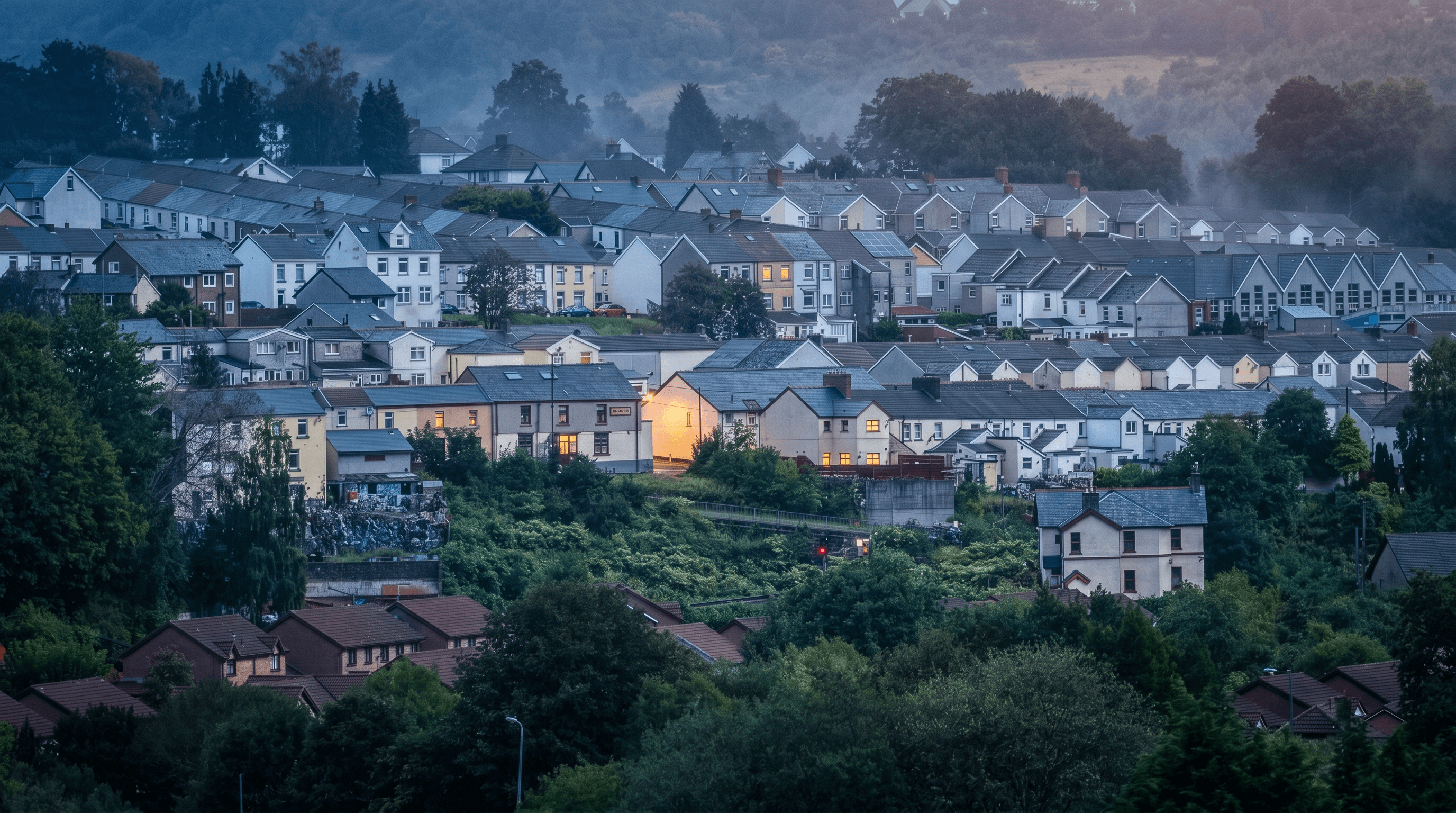 A Merthyr valleys town at dusk — terraced streets on the hillside, one home glowing warm, mist over the hills behind