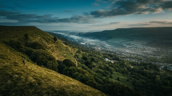 A lone figure on a hillside looking out over a Welsh valley town below, under a moody cinematic sky